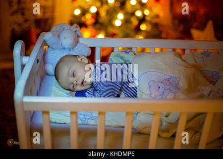 Adorable bébé nouveau-né garçon, dormant dans un lit bébé dans la nuit. Petit garçon au pyjama rayé bleu faire la sieste dans une pièce sombre, la décoration de Noël dans la chambre, Banque D'Images