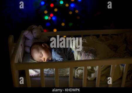 Adorable bébé nouveau-né garçon, dormant dans un lit bébé dans la nuit. Petit garçon au pyjama rayé bleu faire la sieste dans une pièce sombre, la décoration de Noël dans la chambre, Banque D'Images