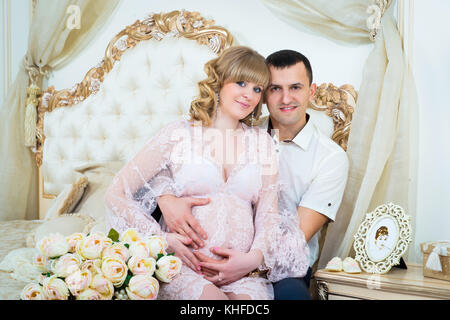 Beau couple, jeune femme enceinte et l'homme serrant avec amour baisers, assis sur le lit, dans un home intérieur Banque D'Images