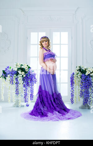 Une happy pregnant woman in a purple jupe longue pose en studio sur un fond de fleurs. Banque D'Images