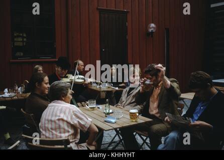 Une photographie qui montre une famille autour d'une table dans un pub, le père divertit les femmes de la famille en buvant sa bière vigoureusement, le fils lit un journal tandis qu'un homme jouant du violon joue de la musique pour eux, d'autres groupes de personnes peuvent être vus dans le fond du patio, 1961. Banque D'Images