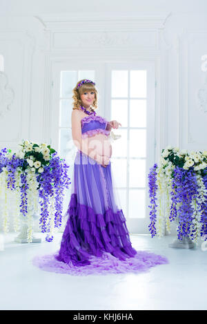 Une happy pregnant woman in a purple jupe longue pose en studio sur un fond de fleurs. Banque D'Images