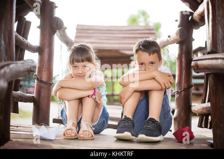 Deux jeunes enfants de race blanche assis à côté de l'autre avec les genoux pliés dans une maison en bois, l'extérieur dès les beaux jours d'été. Ils ont l'air innocent Banque D'Images