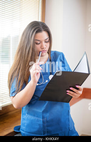 Jeune femme médecin hispaniques en uniforme bleu de l'analyse d'un cas clinique, à la recherche dans un dossier permanent, dans un hôpital près d'une fenêtre, de penser et d'h Banque D'Images