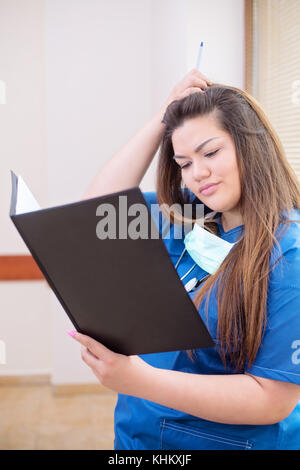 Photo en gros plan de la confondre femme médecin la lecture d'un rapport médical, debout dans un couloir de l'hôpital, le port d'uniforme bleu, r Banque D'Images