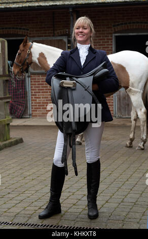 Jolie femme blonde tenant une selle en cuir noir avec son cheval Skewbald. Dans un steableyard. Novembre 2017 Banque D'Images