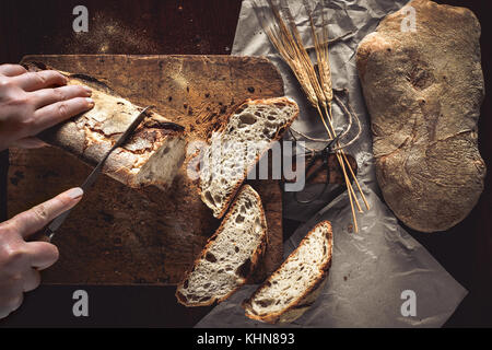 Femme coupe les mains une miche de pain sur la planche en bois rustique et des feuilles de papier, avec les épis de blé et de couteau, vue d'en haut. Banque D'Images