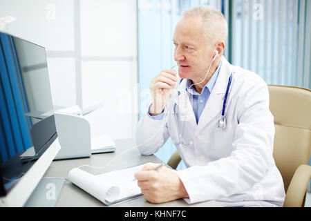 Médecin contemporain des écouteurs consulting les patients grâce à l'appel vidéo alors qu'il était assis en face de l'écran de l'ordinateur par lieu de travail Banque D'Images