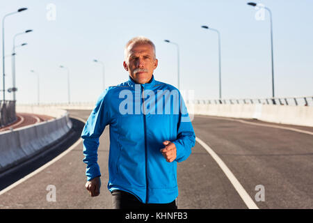 Homme senior actif en veste de sport bleue regardant l'appareil photo pendant la course Banque D'Images