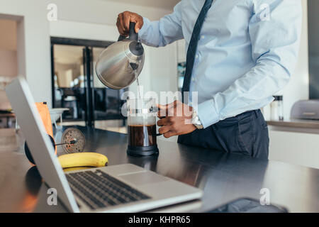 Homme versant de l'eau chaude dans la cafetière. homme d'affaires préparant le café sur la table du petit déjeuner avec un ordinateur portable à côté. Banque D'Images