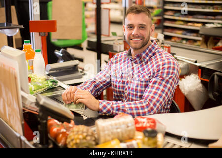 Portrait de beau jeune homme travaillant à caisse de supermarché, posing looking at camera et souriant joyeusement Banque D'Images