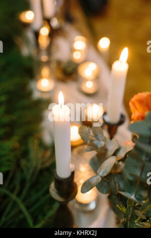 Décoration de mariage, confort, concept. close up de brûler des bougies hautes dans les supports qui sont placés à proximité de roses avec brunch nuance d'orange chaud, sur le blured contexte il y a beaucoup d'éclairage Banque D'Images