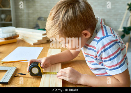 Side view portrait of cute little boy travail du bois assis à table et de prendre des mesures rendant le modèle en bois Banque D'Images