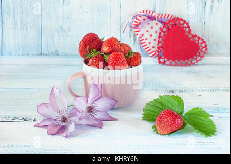 Deux coeurs de tissu avec une fraise mûre dans une tasse en céramique rose cadeau pour la Saint-Valentin sur un fond en bois bleu avec des fleurs délicates clemati Banque D'Images