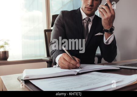 homme d'affaires parlant sur le téléphone portable et prenant des notes. Image recadrée d'un homme assis à son bureau écrivant dans la laiterie et discutant au téléphone. Banque D'Images