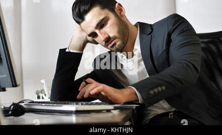 Assez ennuyé jeune homme assis à son bureau devant son ordinateur avec son menton reposant sur sa main, sleepy Banque D'Images
