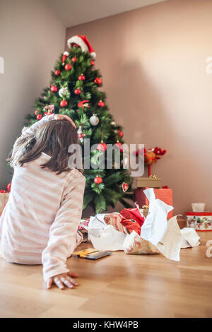 Petite fille ouvrant ses cadeaux sous l'arbre de Noël tôt le matin.Elle est éclairée par la lumière naturelle du matin Banque D'Images