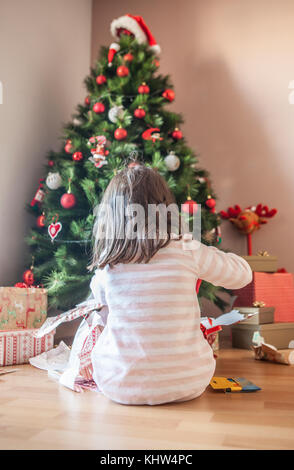 Petite fille ouvrant ses cadeaux sous l'arbre de Noël tôt le matin.Elle est éclairée par la lumière naturelle du matin Banque D'Images