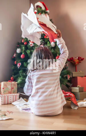 Petite fille ouvrant ses cadeaux sous l'arbre de Noël tôt le matin.Elle est éclairée par la lumière naturelle du matin Banque D'Images