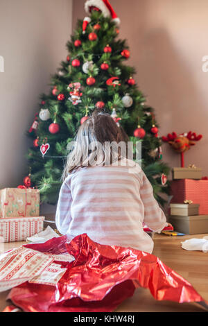 Petite fille ouvrant ses cadeaux sous l'arbre de Noël tôt le matin.Elle est éclairée par la lumière naturelle du matin Banque D'Images