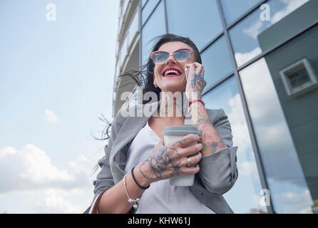 Businesswoman walking outdoors, holding coffee, à l'aide de smartphone, des tatouages sur les mains, low angle view Banque D'Images