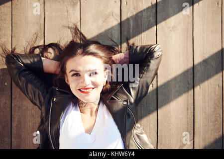 Portrait of young woman lying on decking en bois, les mains derrière la tête, smiling, portrait Banque D'Images