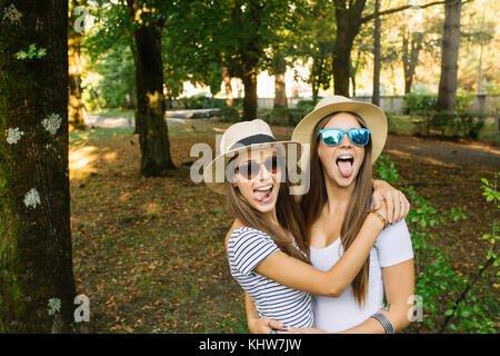 Portrait de deux jeunes amies en chapeau trilby sticking out tongues in park Banque D'Images