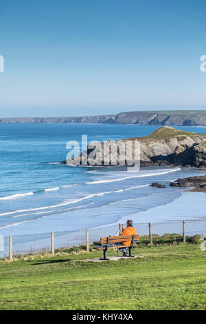 Homme portant une veste rouge vif assise sur un banc surplombant l'île de Porth Head de Trevelgue en arrière-plan. Banque D'Images