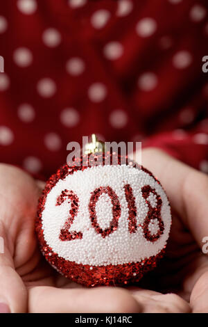 Un gros plan young caucasian femme portant une robe rouge à pois blancs à motifs tenant une boule de noël dans ses mains avec le numéro 2018, comme le nouveau y Banque D'Images