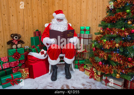 Père Noël assis dans sa grotte entouré par un arbre avec des cadeaux et des boîtes enveloppées cadeau alors qu'il lit son livre préféré : la nuit avant de chri Banque D'Images