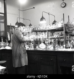 Années 1950, historiques, homme chercheur travaillant dans un laboratoire chemcial avec de nombreuses bouteilles et contenants de verre sur l'établi. Banque D'Images