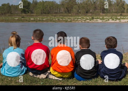 Les enfants en ligne tourne le dos avec chemises de différentes couleurs assise sur le bord de la rivière. Banque D'Images