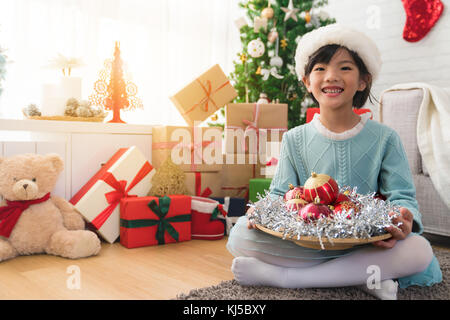 Little cute Asian girl holding a plate of décor rouge shine ball et souriant à l'appareil photo d'une séance sur le plancher dans le salon. Banque D'Images