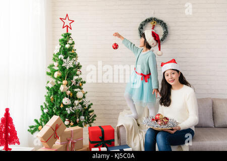 Mère fille asiatique assis et debout sur le canapé décorer l'arbre de Noël à l'intérieur. joyeux noël et de bonnes vacances, le matin avant Noël. Banque D'Images