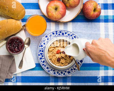 Accueil Petit déjeuner avec des céréales, du lait, des fruits et du pain servi sur bleu et blanc table cloth. woman's hand pouring milk Banque D'Images