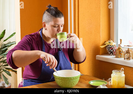 Belle jeune femme aux cheveux courts assis dans la cuisine derrière une table en bois, un mélange les ingrédients dans le bol vert tout en sirotant un thé de gree Banque D'Images