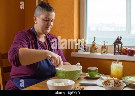 Jeune femme aux cheveux courts portant un tablier bleu, assis dans la cuisine derrière une table, le mélange des ingrédients dans le bol vert, noël cuisson concept Banque D'Images