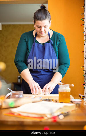 Belle Jeune femme debout dans la cuisine derrière une table de cuisine, pétrir la pâte sur un plan fariné en bois avec ses mains, elle est à la recherche vers le bas Banque D'Images