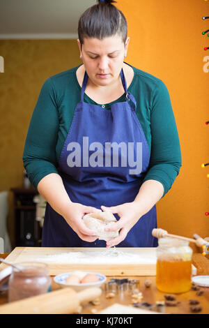 Belle Jeune femme debout dans la cuisine derrière une table de cuisine, pétrir la pâte sur un plan fariné en bois avec ses mains, elle est à la recherche vers le bas Banque D'Images