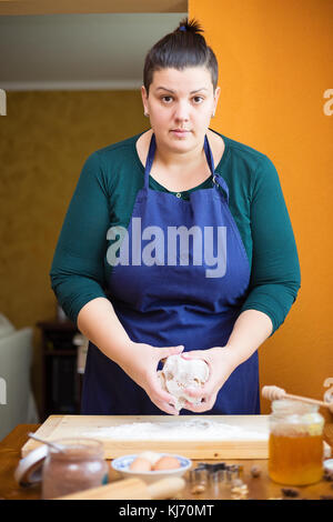 Belle Jeune femme debout dans la cuisine derrière une table de cuisine, pétrir la pâte sur un plan fariné en bois avec ses mains, elle est à la recherche dans Banque D'Images