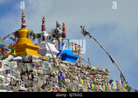 Trois stupas bouddhistes : jaune, blanc et bleu en haut de la maçonnerie, décorés de drapeaux de prière tibetains de couleur. Banque D'Images