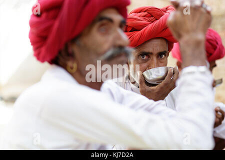 Gros plan d'une homme Rabari avec turban rouge boire d'une coupe d'argent, d'autres hommes sont floues. Le Rajasthan, Inde. Banque D'Images
