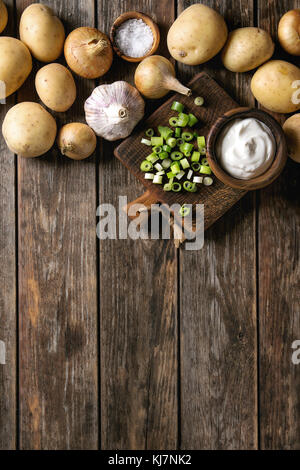 Ingrédients pour cuisiner le dîner. Laver les pommes de terre biologiques entières, l'oignon, l'ail, sel, oignons de printemps et crème fraîche sur la planche à découper en bois ancien Banque D'Images