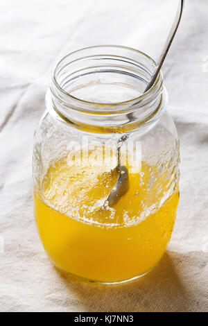 Verre de beurre ghee chaud tiré debout avec une cuillère en bois sur du linge blanc orné sur nappe textile. L'alimentation saine. Close up Banque D'Images