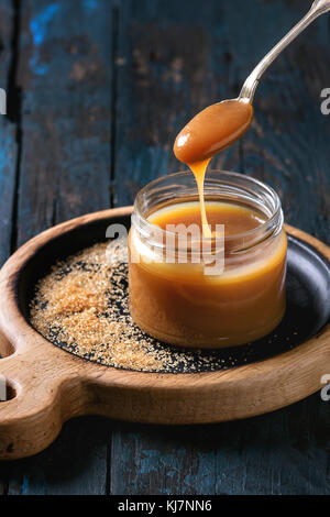 Bocal en verre de sauce caramel salé fait maison en bois servant à bord avec des ingrédients sucre brun ci-dessus. Cuillère de caramel. Plus de vieux bleu foncé Banque D'Images