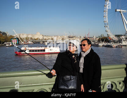 Un couple asiatique selfies posent pour une de Westminster Bridge London England en utilisant un bâton selfies pour tirer le London eye en arrière-plan. Banque D'Images