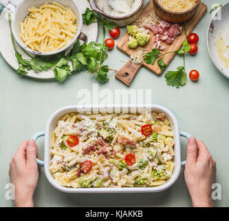 Femme female hands holding casserole de pâtes avec du jambon et le chou romanesco en sauce crémeuse, sur la table de cuisine avec des ingrédients d'arrière-plan, Vue de dessus Banque D'Images