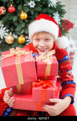 Portrait de gros plan smiling funny little kid holding nombreux présente rouge décorée avec des rubans d'or. petit garçon fêter Noël avec la famille. vert Banque D'Images