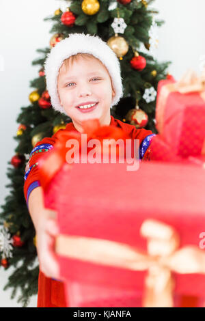 Closeup portrait of happy smiling child holding pile de boîtes présente dans les mains. boy wearing santa hat et red pyjamas. maison de vacances arbre verdoyant backgroun Banque D'Images