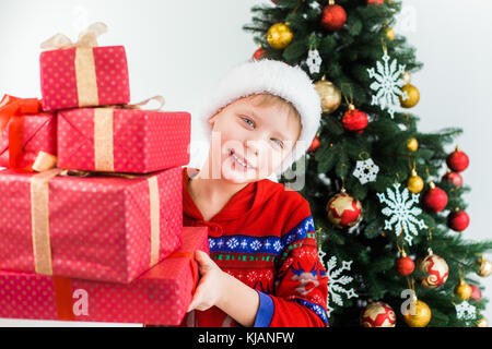 Portrait of cute funny little kid habillés en pyjamas nuit rouge et un chapeau de Père Noël. white happy kid holding pile de vacances présente dans les mains et sourit Banque D'Images